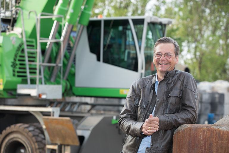 Ein Mann in Lederjacke steht vor einer großen Baumaschine und lächelt in die Kamera.
