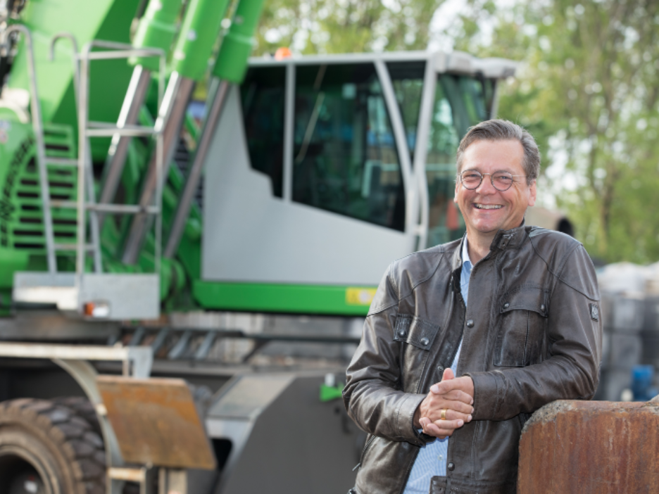Ein Mann in Lederjacke steht vor einer großen Baumaschine und lächelt in die Kamera.