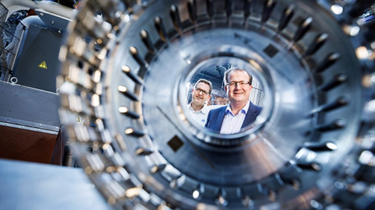 Zwei Männer in einer Fertigungshalle für Metallbauteile, im Vordergrund ein solches Bauteil