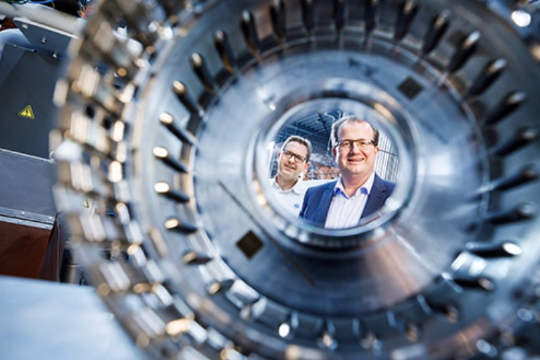 Zwei Männer in einer Fertigungshalle für Metallbauteile, im Vordergrund ein solches Bauteil