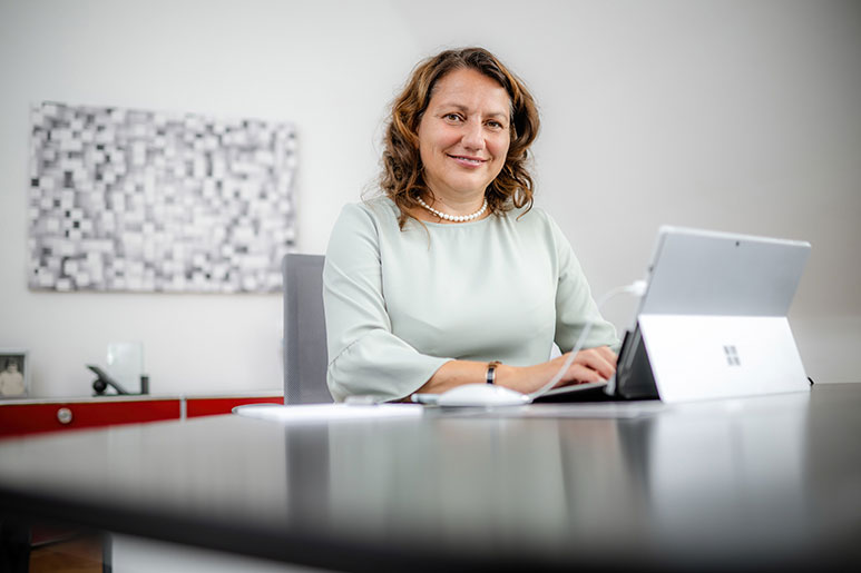 Sabine Hansen sitzt in einem weißen, hellen Büro an ihrem Schreibtisch, tippt etwas in ihr Tablet und schaut in die Kamera.