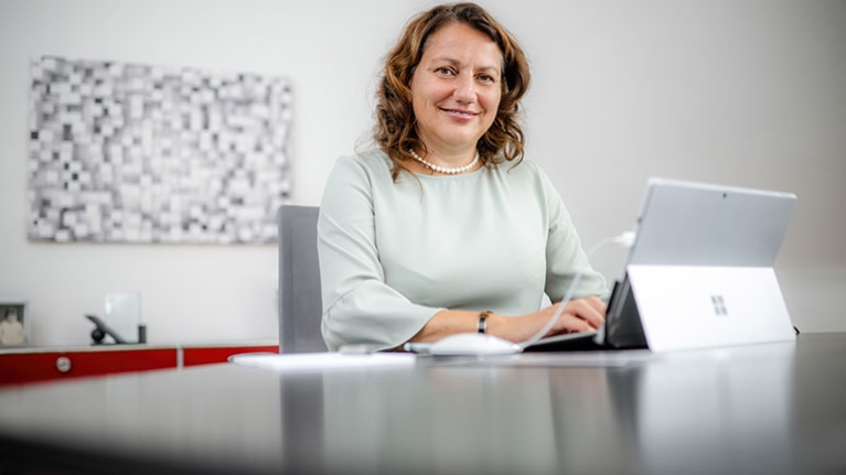 Sabine Hansen sitzt in einem weißen, hellen Büro an ihrem Schreibtisch, tippt etwas in ihr Tablet und schaut in die Kamera.