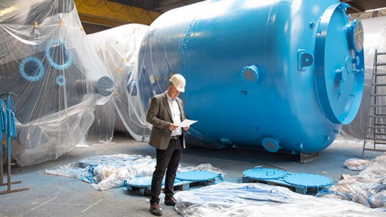 Dr. Jörg Doege, Geschäftsführer von Inprocoat, mit Schutzhelm vor einem Metalltank in einer Werkshalle
