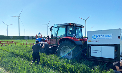Traktor auf einem grünen Feld, davor steht ein Mann, im Hintergrund Windräder