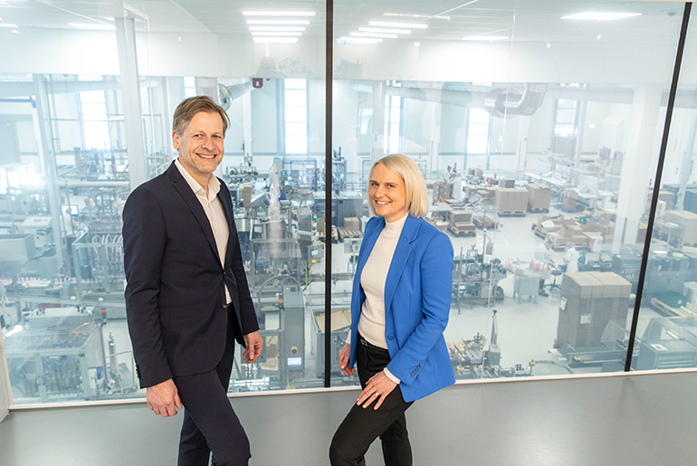 Babor-Geschäftsführer Stefan Kehr war Claudia Brendt von der Förderberatung Rheinland stehen vor einer scheibe, die den Produktionsbereich abtrennt