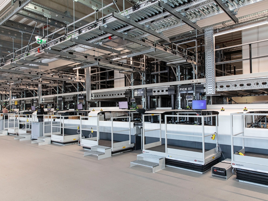 Graue Kästen auf Ketten an meherern Arbeitspositionen für Logistikmitarebeitende, die die Lieferungen zusammenstellen