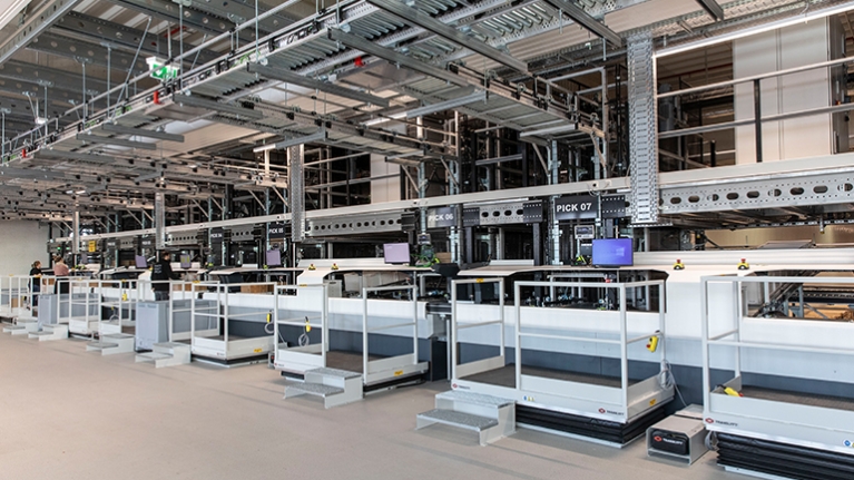 Graue Kästen auf Ketten an meherern Arbeitspositionen für Logistikmitarebeitende, die die Lieferungen zusammenstellen