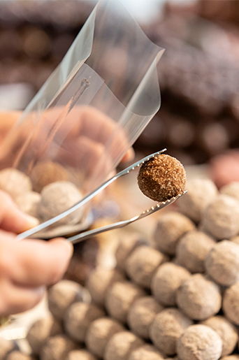 Eine handgemachte Praline wird mit einer Zange in eine Tüte eingepackt. im Hintergrund sind weitere Pralinen zu sehen.