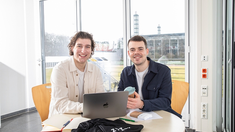 Die zwei GEschäftsführer von Kiman Sport sitzen an einem Laptop und halten Produkte ihrer Sportartikel Firma in den Händen