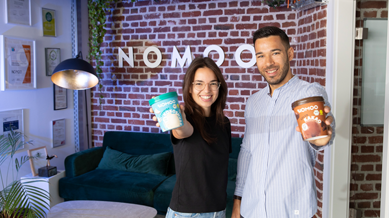 Zwei Personen; eine junge Frau und ien junger Mann, stehen vor einer Backsteinwand und halte zei Eisbecher in der Hand