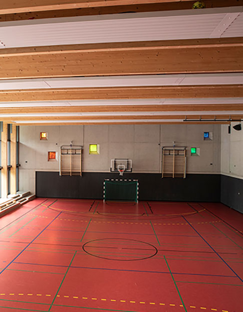 Innenraum einer Schul-Sporthalle mit Basketballkorb, roten Bodenmarkierungen und großen Fenstern.