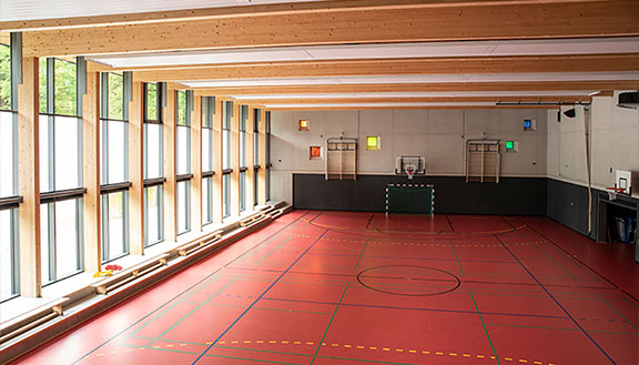 Innenraum einer Schul-Sporthalle mit Basketballkorb, roten Bodenmarkierungen und großen Fenstern.