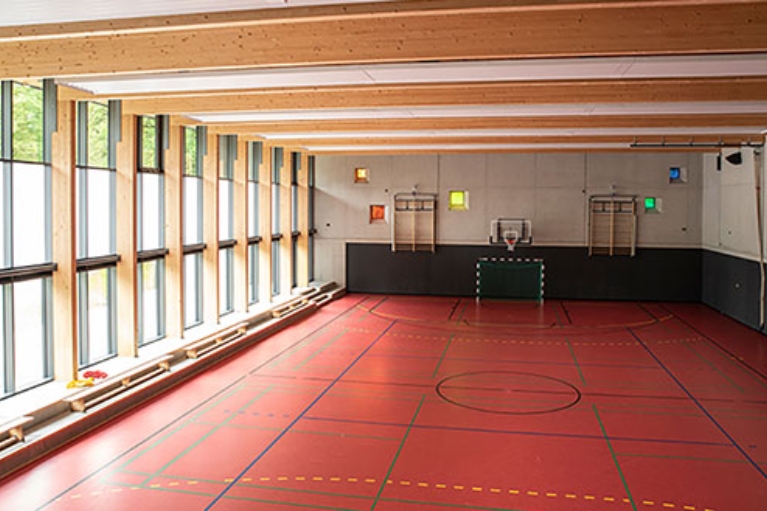 Innenraum einer Schul-Sporthalle mit Basketballkorb, roten Bodenmarkierungen und großen Fenstern.