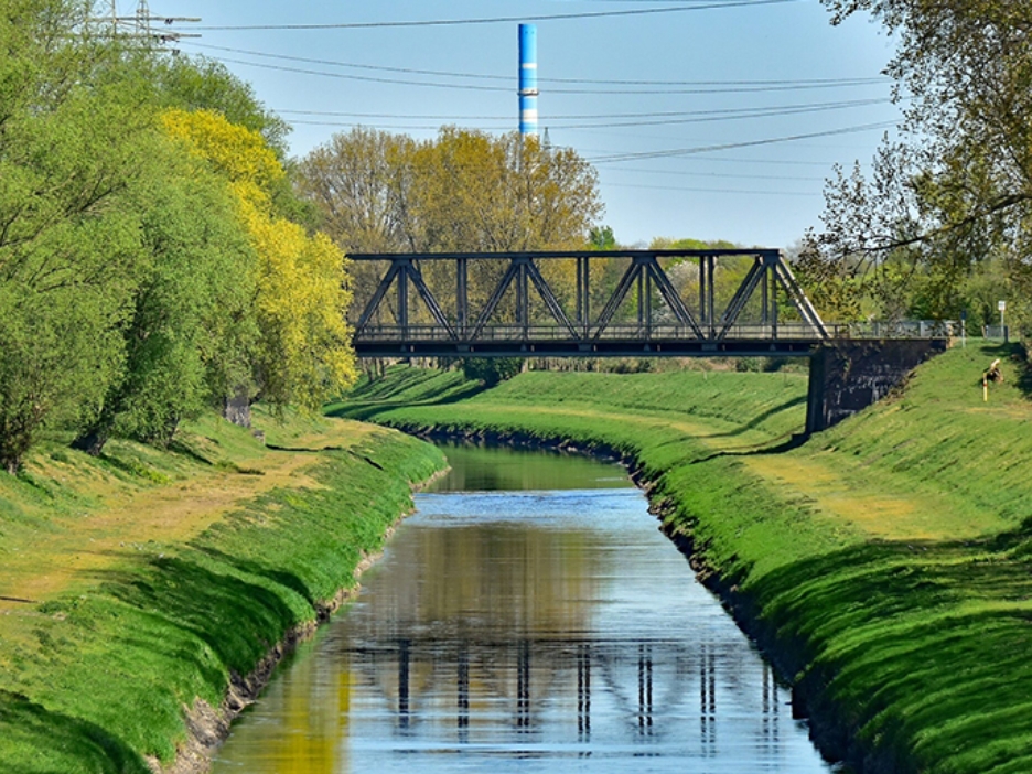 Geradliniger Fluss mit steilen Deichen eingefasst und bewachsener Prommenaden und überspannt von alter Eisenbahnbrücke