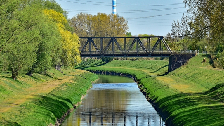 Geradliniger Fluss mit steilen Deichen eingefasst und bewachsener Prommenaden und überspannt von alter Eisenbahnbrücke