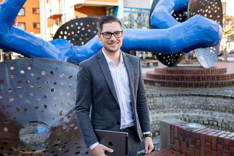 Ein Mann vor einem großen, modernen Kunstobjekt in einer Fußgängerzone. In der Hand hat er ein iPad.