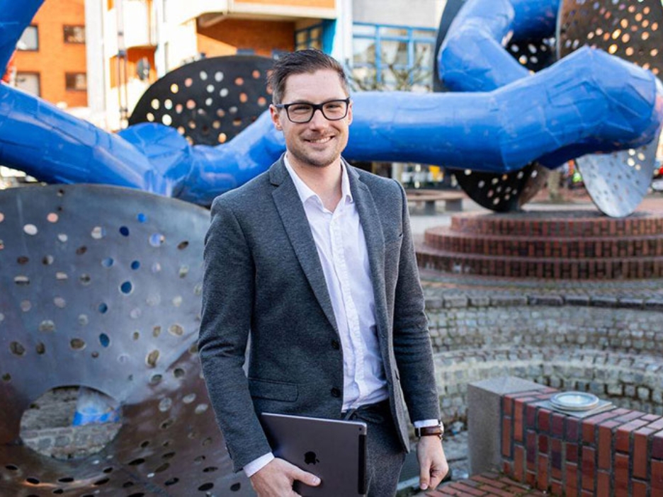 Ein Mann vor einem großen, modernen Kunstobjekt in einer Fußgängerzone. In der Hand hat er ein iPad.