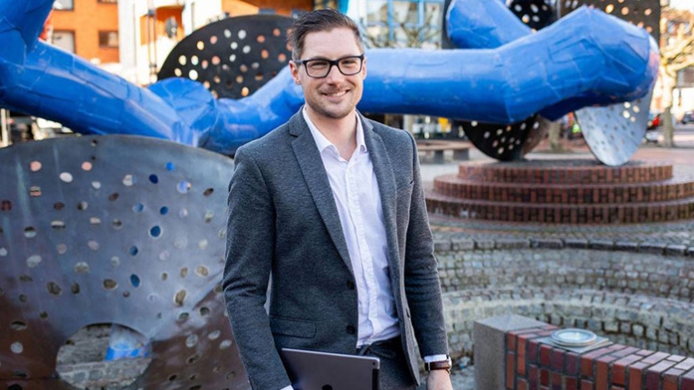 Ein Mann vor einem großen, modernen Kunstobjekt in einer Fußgängerzone. In der Hand hat er ein iPad.