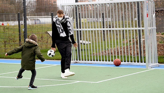 Ein Junge spielt mit Manuel Neuer auf einem Kunstrasenplatz Fußball.