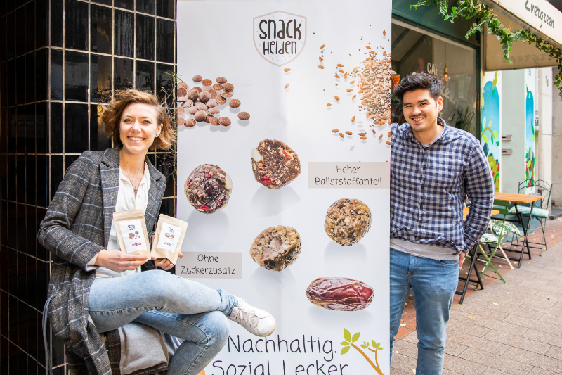 Kerstin Drazkiewicz und David Herzmann von den Snackhelden lächelnd vor einem Werbebanner ihrer Produkte