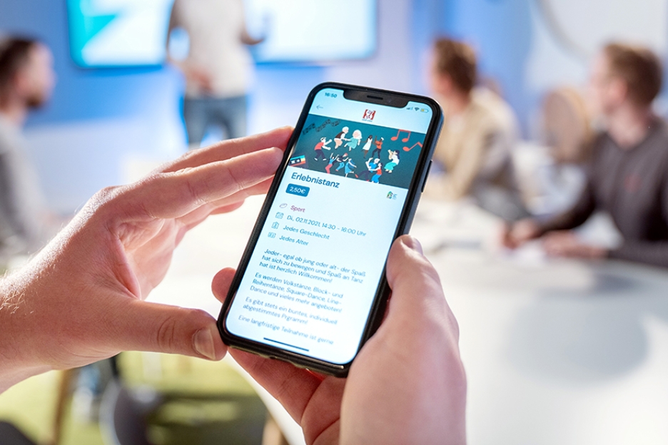 Das Foto zeigt Hände, die ein Smartphone halten, auf dem die Oberfläche der App Tremaze zu finden ist. Im Hintergrund sind vier Männer zu sehen, die ein Meeting abhalten.