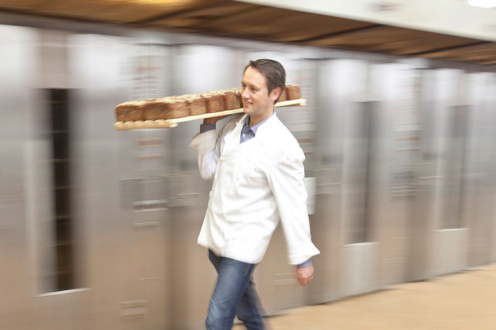 Bäcker Roland Schüren trägt ein großes Blech mit Broten. Im Hintergrund verschwommen große Backöfen