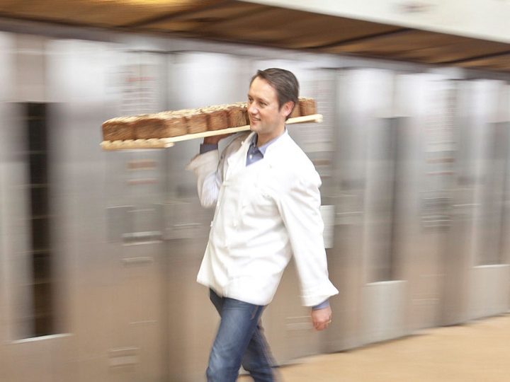 Bäcker Roland Schüren trägt ein großes Blech mit Broten. Im Hintergrund verschwommen große Backöfen