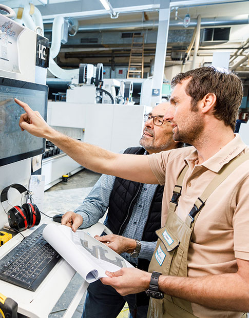 Zwei Männer in einer Werkshalle vor einer modernen Maschine. Einer deutet auf den Bildschirm der Maschine.