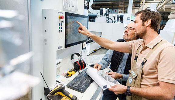 Zwei Männer in einer Werkshalle vor einer modernen Maschine. Einer deutet auf den Bildschirm der Maschine.