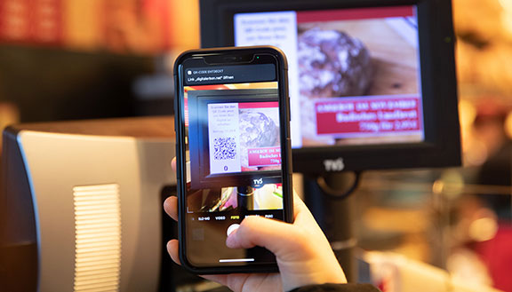 Bildschirm einer modernen Kasse, davor ein Smartphone, das den QR-Code auf dem Bildschirm scannt.
