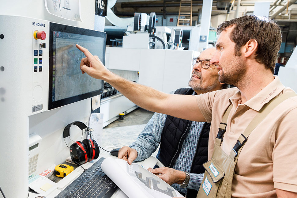Zwei Männer in einer Werkshalle vor einer modernen Maschine. Einer deutet auf den Bildschirm der Maschine.