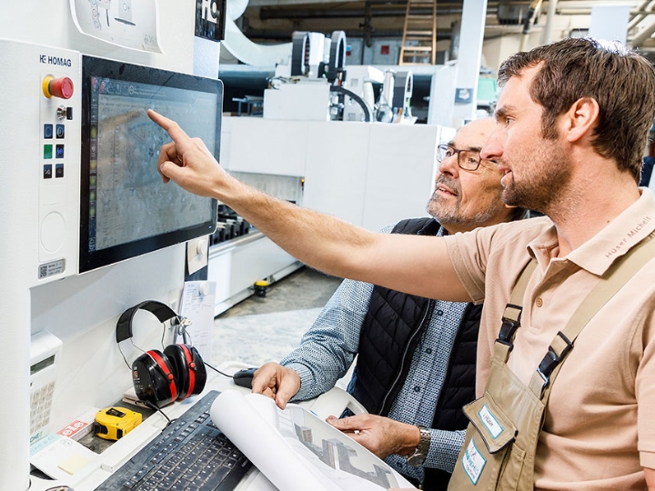 Zwei Männer in einer Werkshalle vor einer modernen Maschine. Einer deutet auf den Bildschirm der Maschine.