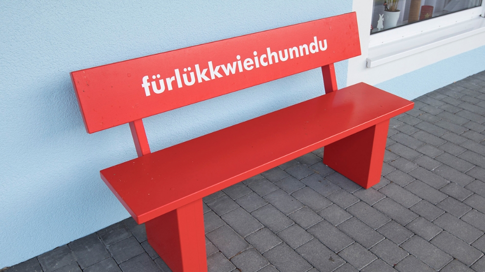 Rote Holzbank vor einem Haus. Auf der Bank die Aufschrift Für Lück wie ich unn du