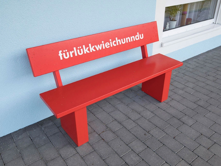 Rote Holzbank vor einem Haus. Auf der Bank die Aufschrift Für Lück wie ich unn du