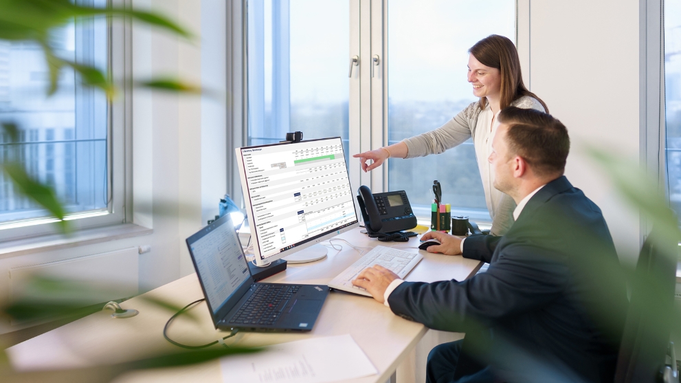Eine Frau und ein Mann an einem Schreibtisch im Büro. Die Frau deutet mit dem Finger auf eine Tabelle auf dem Monitor.
