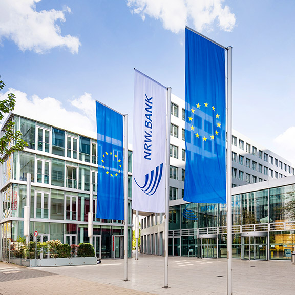 Das Bild zeigt die Fahne der NRW.BANK in der Mitte von zwei EU-Fahnen vor dem Gebäude. Die NRW.BANK fördert die Internationalisierung von Unternehmen.