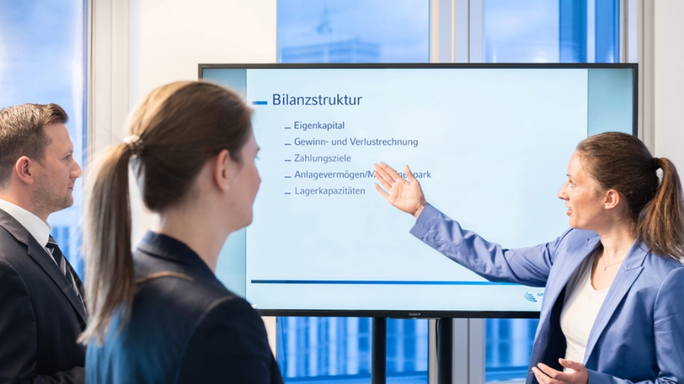 Auf einem Monitor steht das Wort Bilanzstruktur. Eine Frau erklärt etwas dazu.