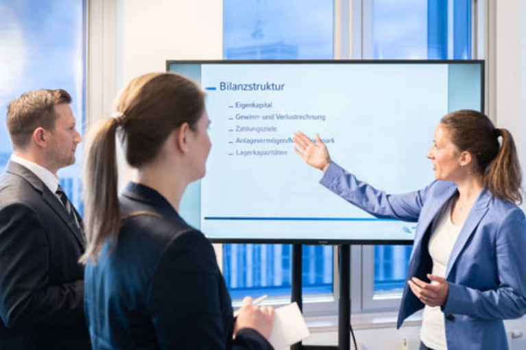 Auf einem Monitor steht das Wort Bilanzstruktur. Eine Frau erklärt etwas dazu.