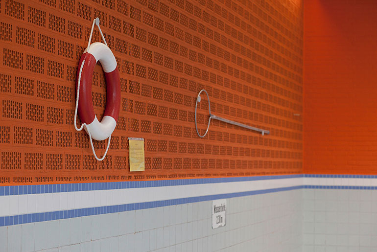 Orangefarbene Wand eines leeren Schwimmbads. An der Wand hängt ein Rettungsring und eine Rettungsstange