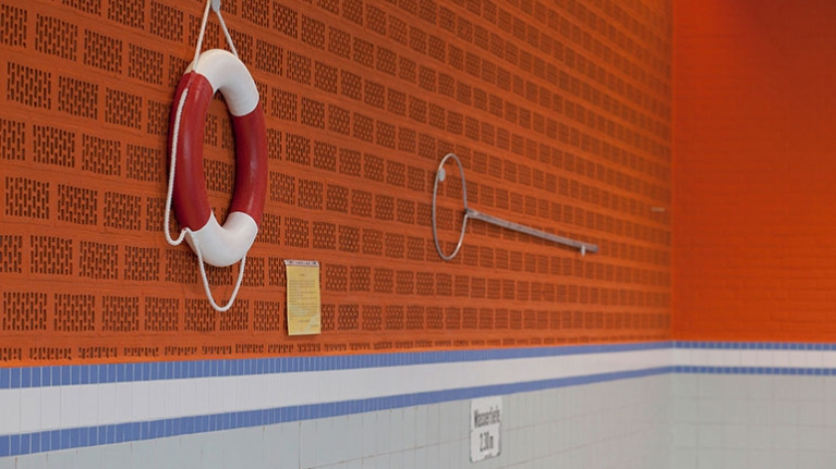 Orangefarbene Wand eines leeren Schwimmbads. An der Wand hängt ein Rettungsring und eine Rettungsstange