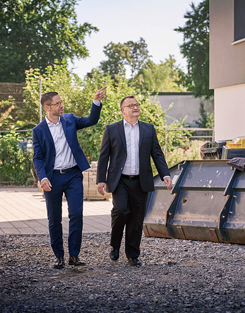 Zwei Männer auf einem Schotterweg vor einem Wohnhaus. Der eine zeigt auf das Haus.