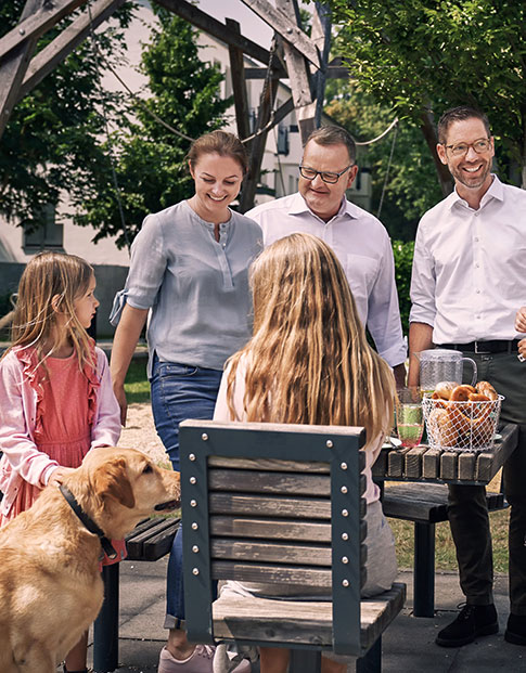 Zwei Männer, eine Frau, zwei Mädchen und ein Hund beim Picknick im Freien an einem Holztisch
