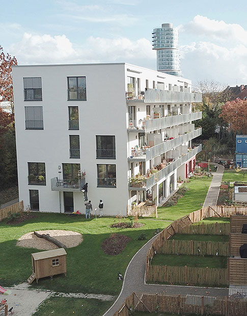 Weißes Mehrfamilienhaus mit Balkonen und einer Gartenanlage. Im Hintergrund ein futuristisches Hochhaus