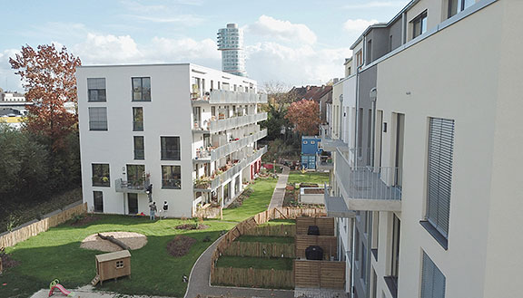 Weißes Mehrfamilienhaus mit Balkonen und einer Gartenanlage. Im Hintergrund ein futuristisches Hochhaus