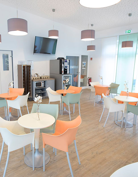 Mehrere Tische mit grünen, weißen und orangenen Stühlen in einer hellen Cafeteria