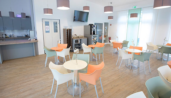Mehrere Tische mit grünen, weißen und orangenen Stühlen in einer hellen Cafeteria