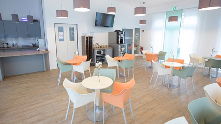 Mehrere Tische mit grünen, weißen und orangenen Stühlen in einer hellen Cafeteria