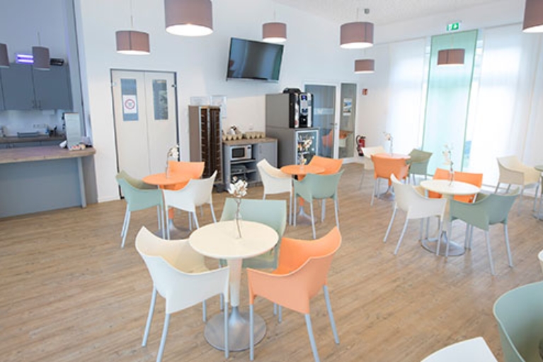 Mehrere Tische mit grünen, weißen und orangenen Stühlen in einer hellen Cafeteria