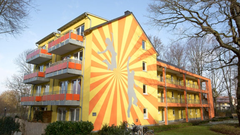 Orange-gelbe Fassade eines Studentenwohnheims mit aufgeklebten Silhouetten von Sportlern