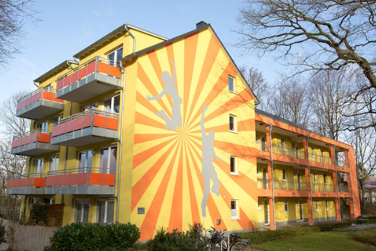 Orange-gelbe Fassade eines Studentenwohnheims mit aufgeklebten Silhouetten von Sportlern